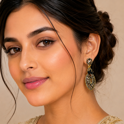 A close-up portrait of a woman wearing a large, ornate gold-toned dangle earring with black and green stone detailing, paired with a beige embroidered traditional outfit against a soft beige background.