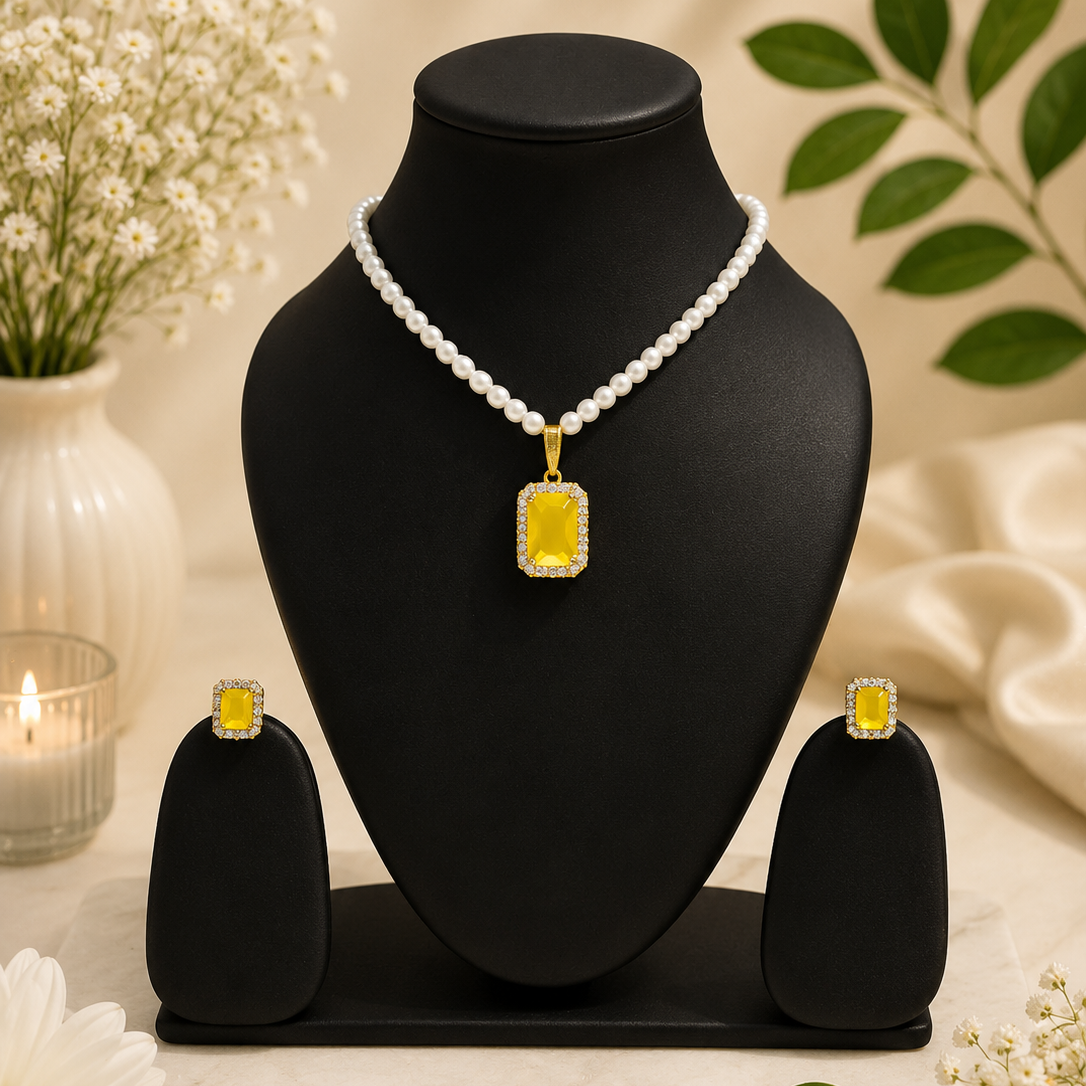 A jewelry set displayed on a black mannequin bust, featuring a pearl-bead necklace with a rectangular yellow gemstone pendant, matching yellow gemstone stud earrings, and a black jewelry box. The set is styled with white flowers, green leaves, and a lit candle on a beige background.