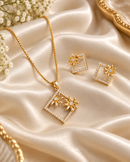 Gold jewelry set featuring a necklace and matching earrings with square diamond-like frames and starburst floral details, arranged on draped champagne satin fabric with small white flowers.