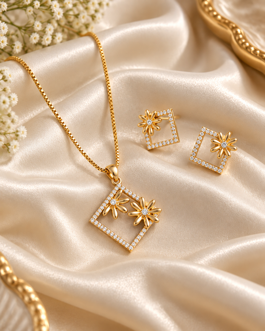Gold jewelry set featuring a necklace and matching earrings with square diamond-like frames and starburst floral details, arranged on draped champagne satin fabric with small white flowers.